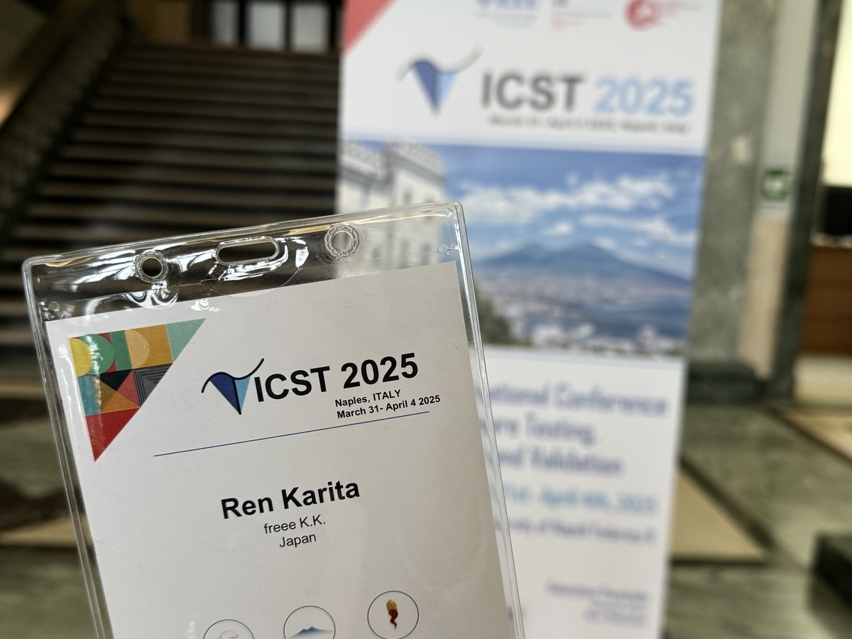 ICST 2025カンファレンスの参加者に配られた名札。名札には参加者名とfreee K.K.、Japanの所属情報が記載されており、背景にはカンファレンスのポスターやバナーが写っている。