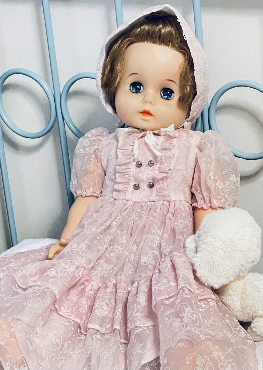 ソフビ製の抱き人形。ピンクのレースのドレスを着て、同じ色の帽子を被っている。白い犬のぬいぐるみが懐いている。