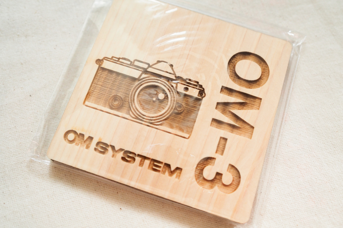 OM SYSTEMブースで配布された木製のOM-3カメラ型コースター