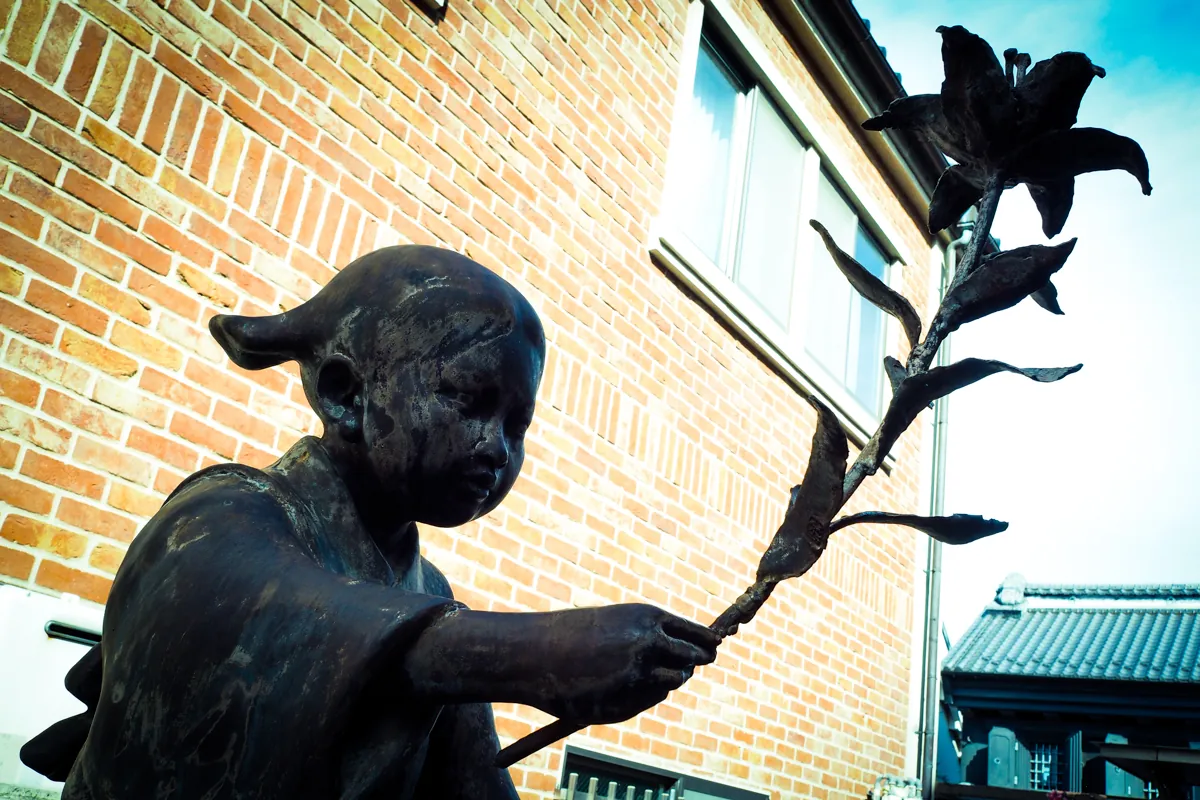 川越まつり会館横に立つ、佐藤健次郎氏作の『まごころ像』。レンガの壁を背景に百合の花を持つ少女の像が、柔らかな光と影の中で静かに佇む。撮影者の心を惹きつけ、一度通り過ぎた後も引き返して撮影した、温かみのある情景。