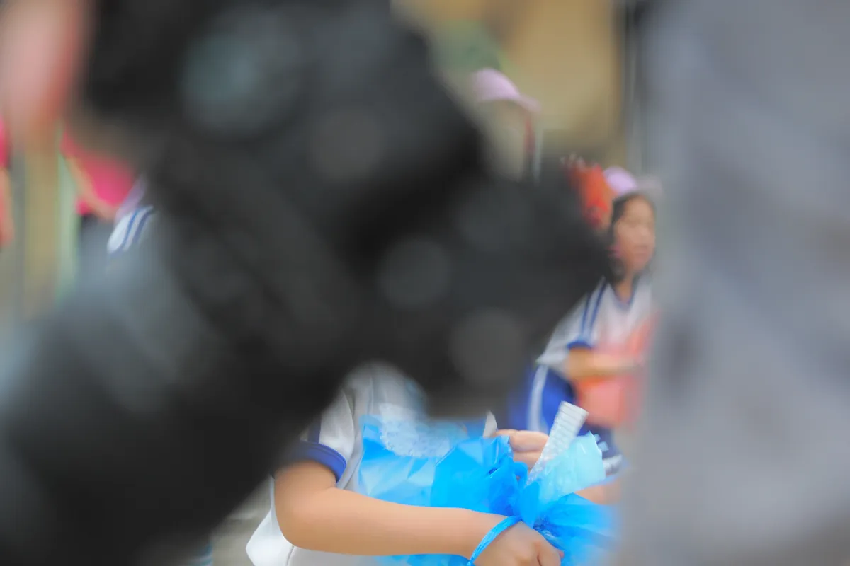 大きな黒いカメラレンズが手前にぼやけて写り、その奥に青いポンポンを持つ体操服姿の子供が立っている写真