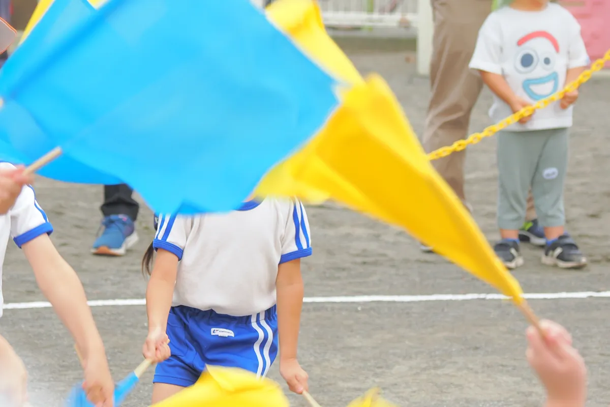 運動会で青と黄色の旗を両手に持ち、振っている体操服姿の子供の全身像