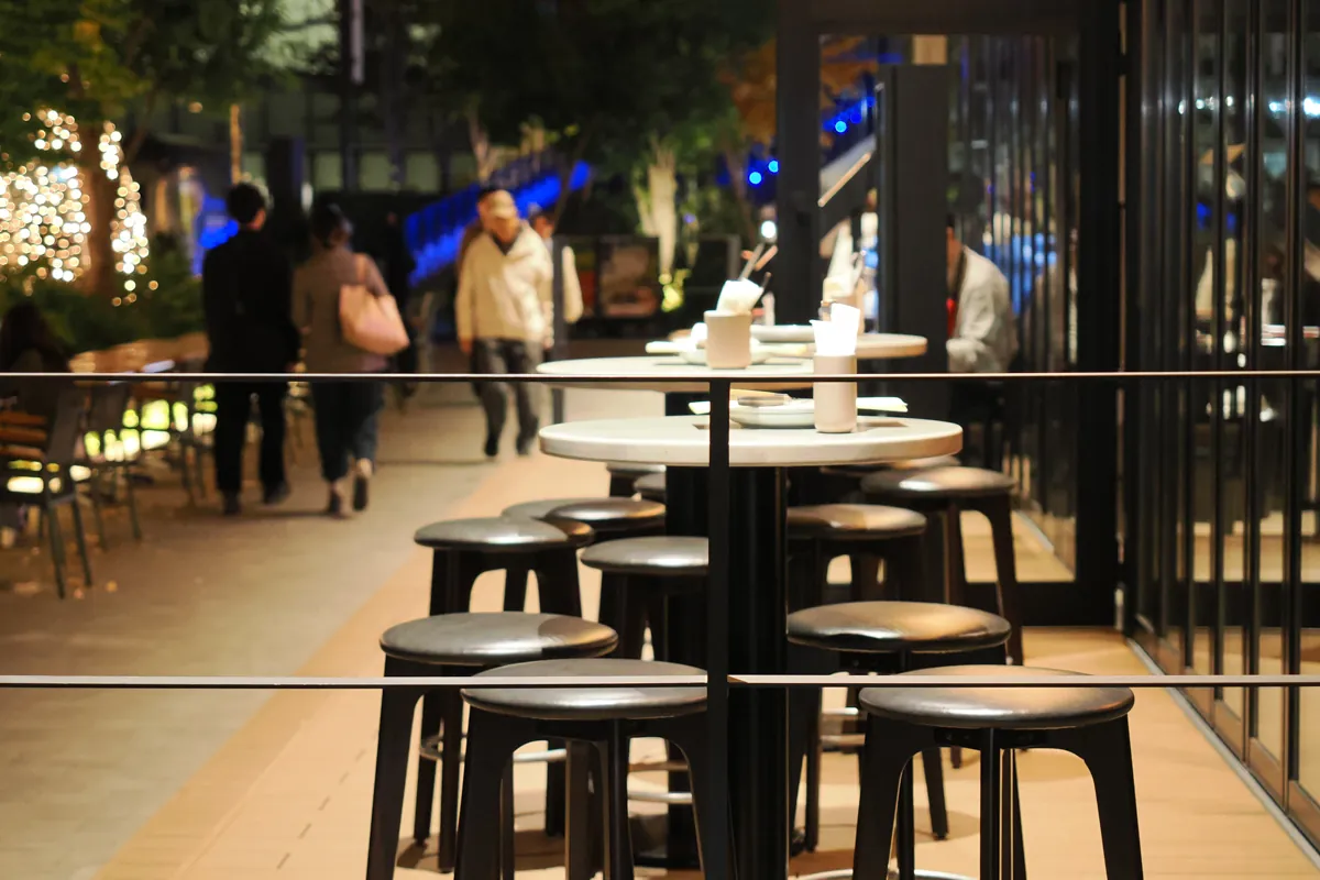 立川グリーンスプリングスの夜のカフェテラス。手前には空のテーブルと椅子が並び、奥にはぼやけた人影とイルミネーションの光が見える。