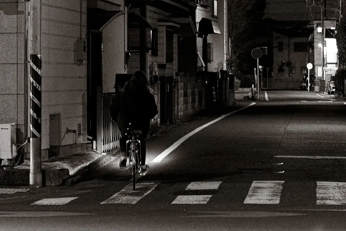 夜のモノクロームスナップ。街灯に照らされたアスファルトの白線の上を、自転車に乗った人物が奥へ走り去る背中。中望遠レンズ特有の圧縮効果と、夜の静寂を感じさせる粒子感のある描写。