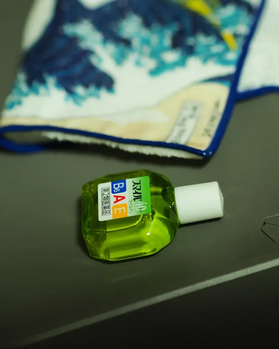 デスクの上に置かれた緑色の目薬「スマイル40EX」の接写。背景には葛飾北斎の『神奈川沖浪裏』がプリントされた青い縁取りの白い眼鏡ふきが柔らかくボケて写っている。