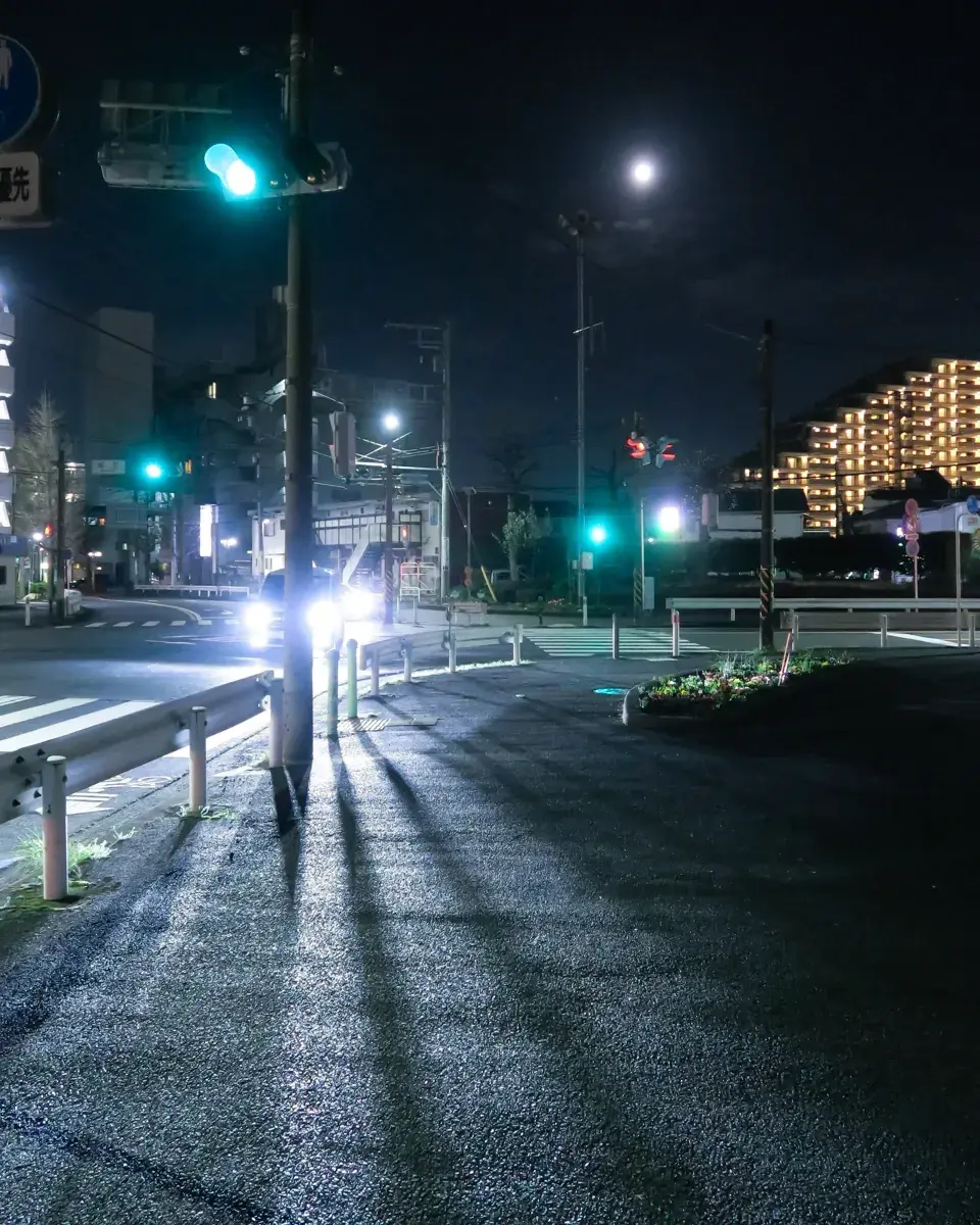深夜の交差点。車のヘッドライトが電柱の影を鋭くアスファルトに叩きつけている。奥に月。