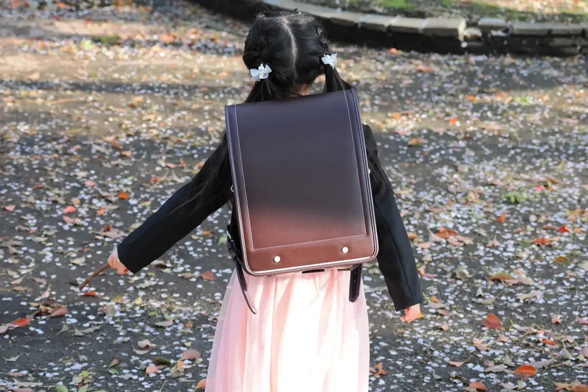 茶色のランドセルを背負った女の子の後ろ姿。散った桜の花びらが地面を埋めている。