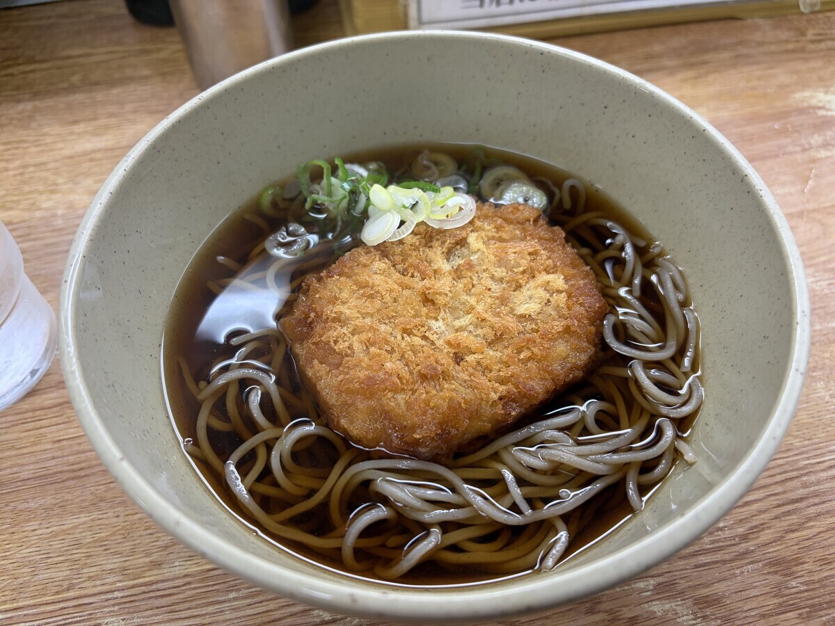 和風の陶器の丼に盛り付けられた温かいそばの料理が写っている。つゆは濃いめの色をしており、中にはそば麺が入っている。中央には、衣がサクサクとした見た目の揚げ物(おそらくコロッケ)が乗せられている。揚げ物の上や周囲には刻んだ青ねぎと白ねぎがトッピングされている。背景には木製のテーブルが写っており、左側には白いプラスチック製のコップが見える。