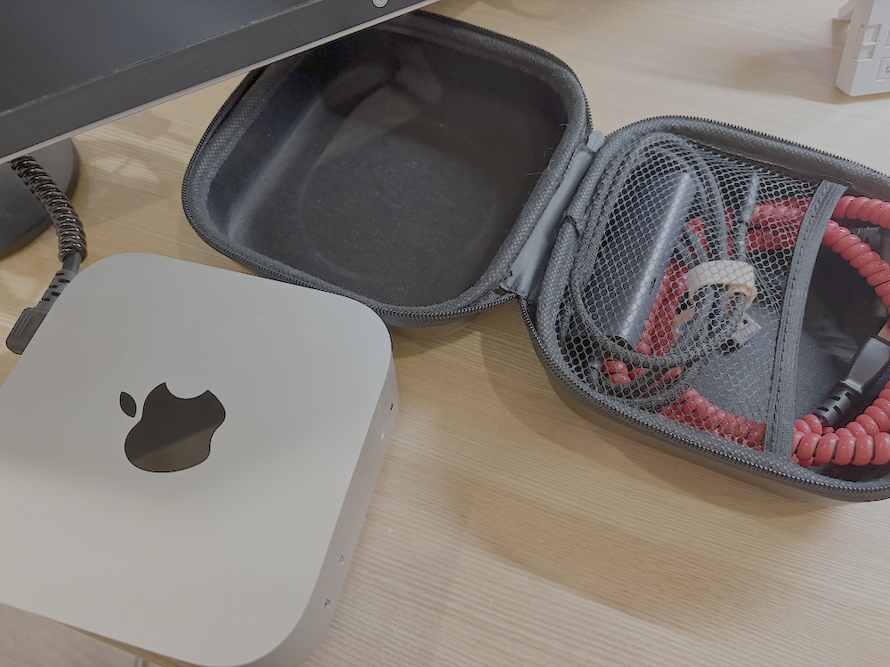 ALT説明:  木製のデスクの上に、いくつかの電子機器が置かれている。左側にはAppleのロゴが入ったシルバーのMac miniがあり、その後方にはモニターの一部と接続ケーブルが見えている。右側には、開いた黒いハードケースがあり、右側のポケット部分に赤いコイル状のケーブル、USBケーブル、アダプターなどが収納されている。ケースの左側は空で、クッション素材が内張りされている。