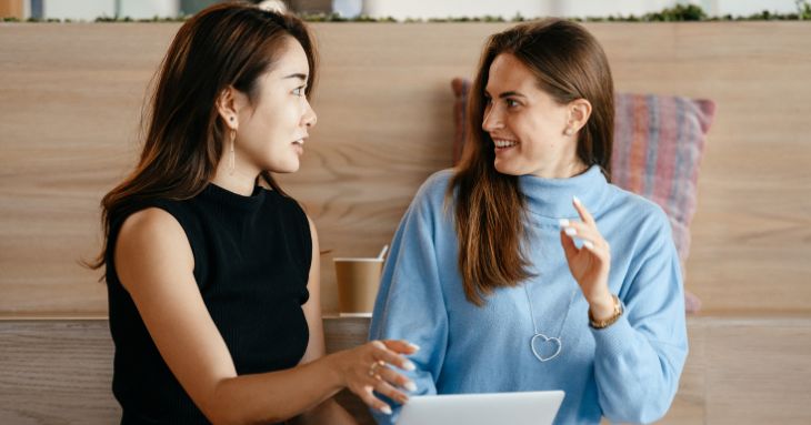 女性二人が並んで座り、一方が話しているのをもう一方がほほえみながら聞いている
