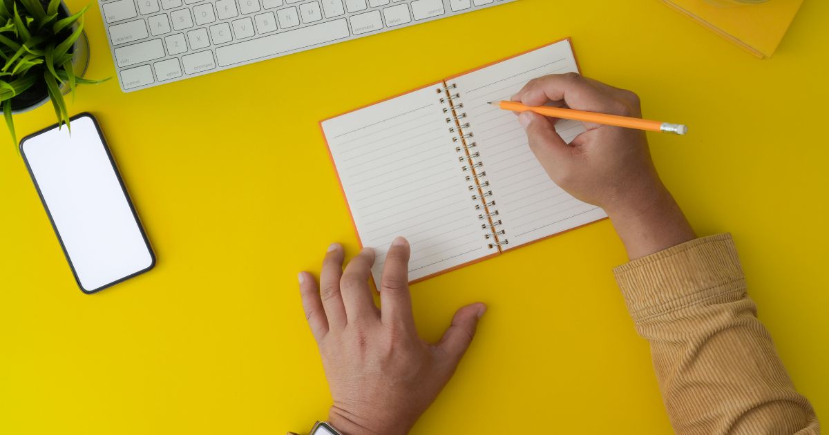 黄色いデスク上で、えんぴつでメモ帳に記入しようとする人の手元