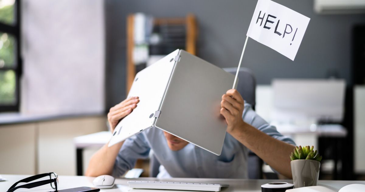 オフィスのデスクで、頭上にノートパソコンをかざして身を守りながら「HELP!」と書かれた小さな旗を掲げ、助けを求めているビジネスパーソンの様子