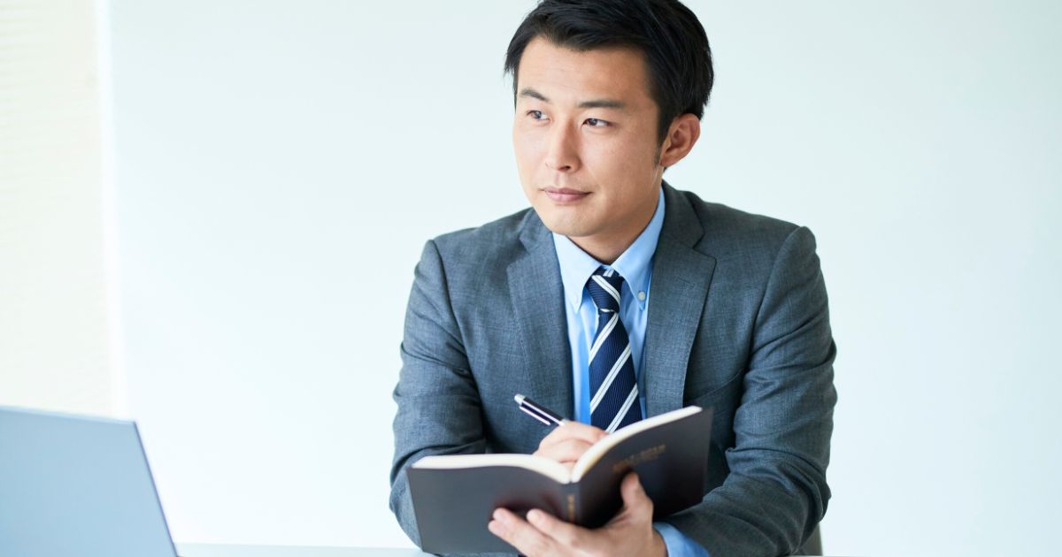 スーツ姿の男性がノートにペンを走らせながら考え込む。デスクにはノートPCが置かれている