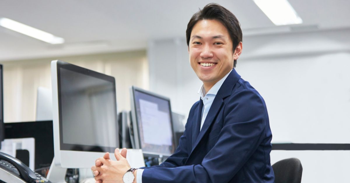 明るいオフィスでデスクに座り、ディスプレイの前で笑顔を向けるネイビーのスーツ男性