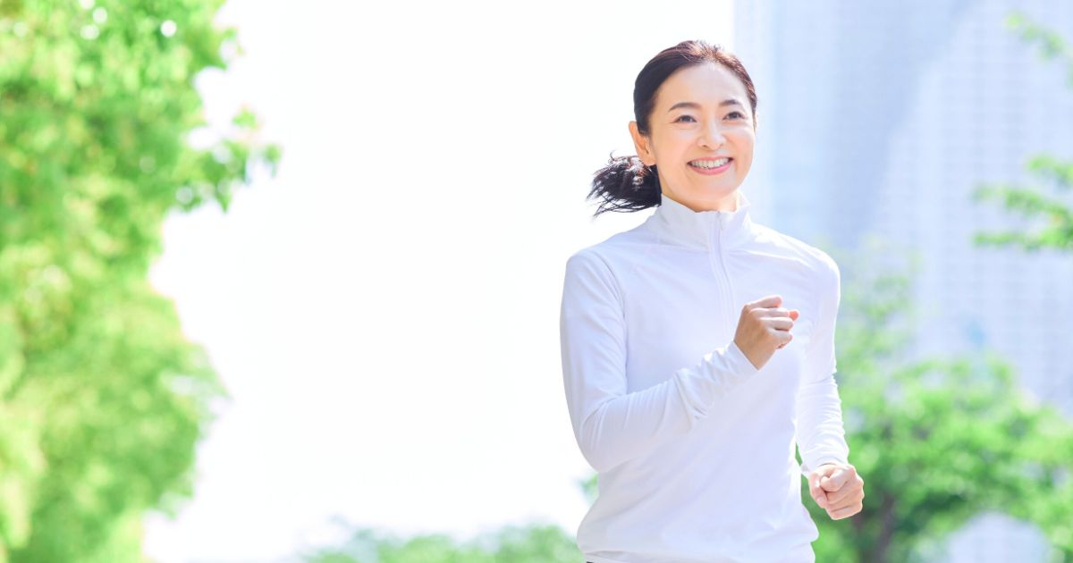木々の緑の中、白いウエアで朝日を浴びながら軽くジョギングする女性