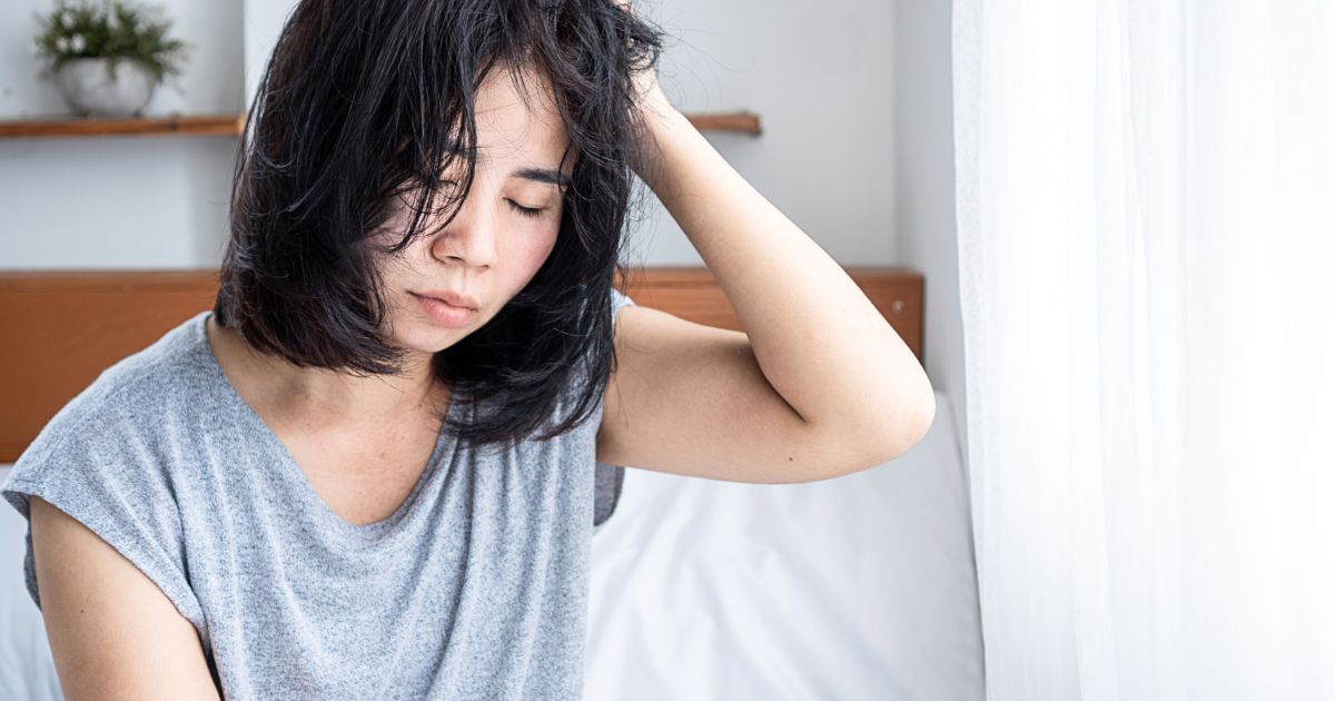 寝起きのだるさで目を閉じ、ベッドの上で髪を押さえる女性