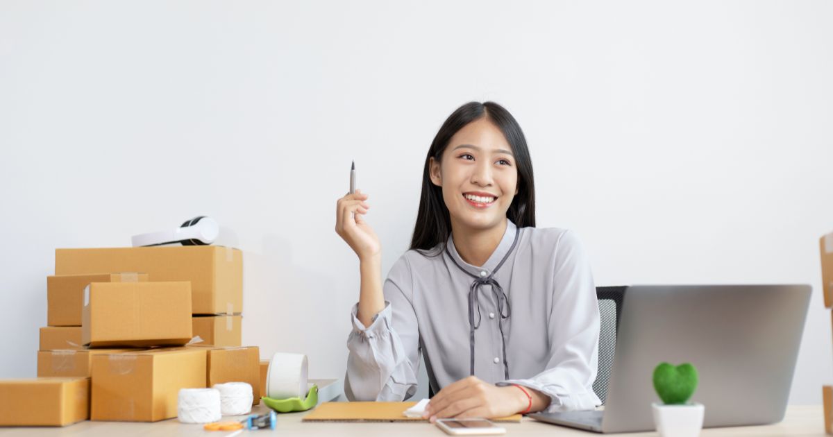 オフィスで笑顔を見せるビジネスパーソン。オンライン業務や発送準備をしながらアイデアを考えている。