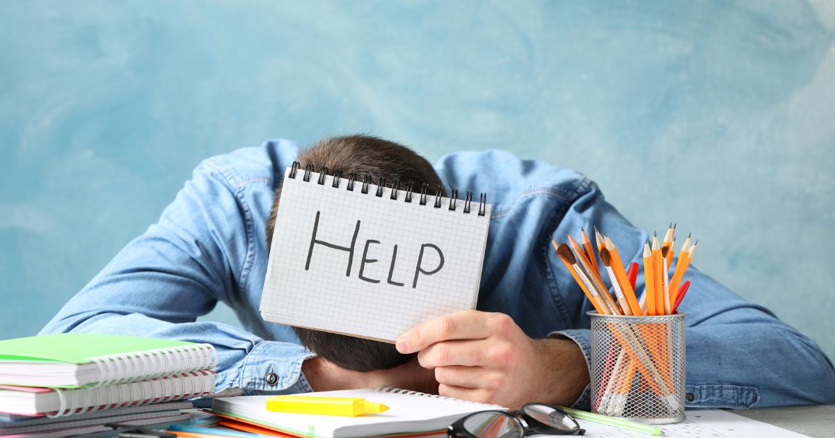 デスクにうつ伏せになり、ノートに「HELP」と書いて助けを求める男性。