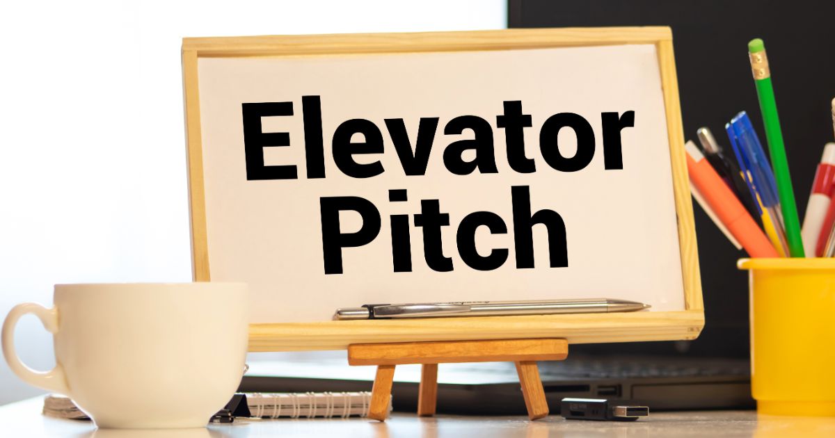 『Elevator Pitch』と書かれたボードが小さなイーゼルに立てかけられている。手前には白いコーヒーカップとノート、背景には色とりどりのペン立てが置かれたデスクが見える