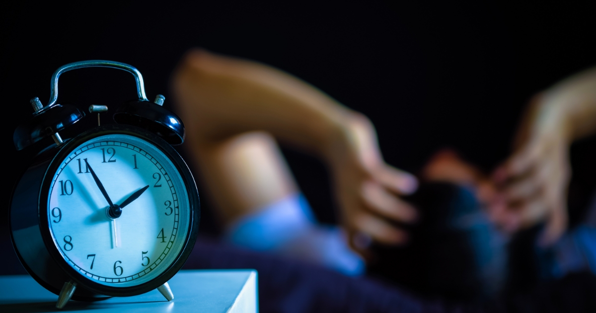ベッドサイドに置かれた目覚まし時計と、背景で横になって眠る人物
