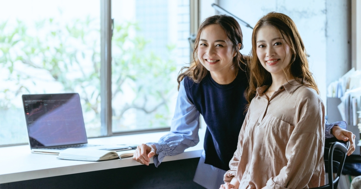 明るい自然光が差し込むワークスペースで、デスク横に並んで微笑む2人の女性。ノートPCとノートが置かれた机が見える