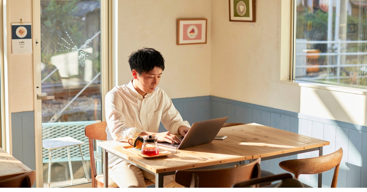 日差しの入るカフェで、木のテーブルに座りノートPCで作業する男性