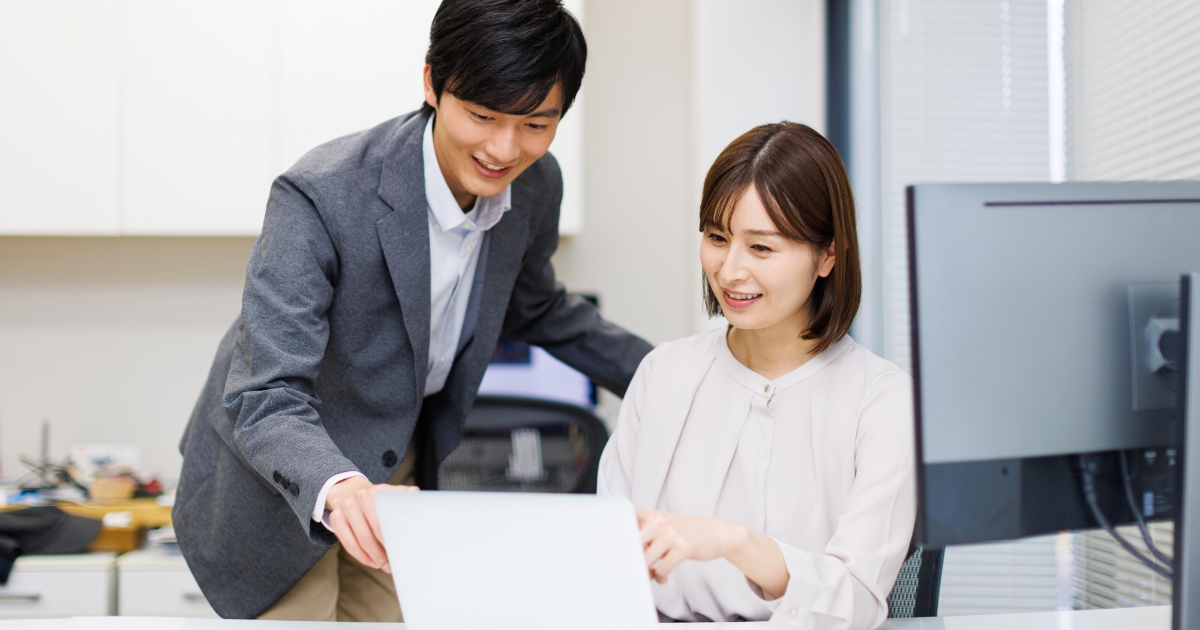 オフィスで、スーツ姿の男性が座っている女性のノートPC画面を指し示しながら説明している様子
