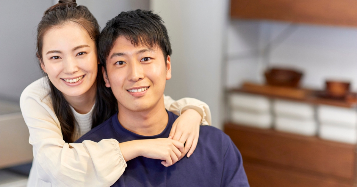 リビングで笑顔の男女が寄り添い、女性が男性の肩に腕を回している様子