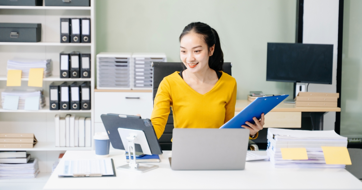 黄色のトップスの女性が、オフィスのデスクでノートPCとタブレットを使いながら青いバインダーを手に仕事をしている
