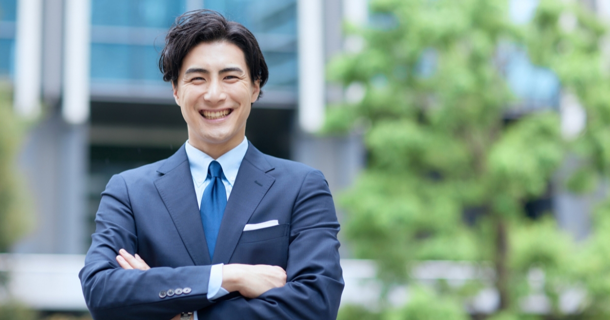 スーツ姿の男性が腕を組み、ビルと緑を背景に屋外で笑顔で立つ