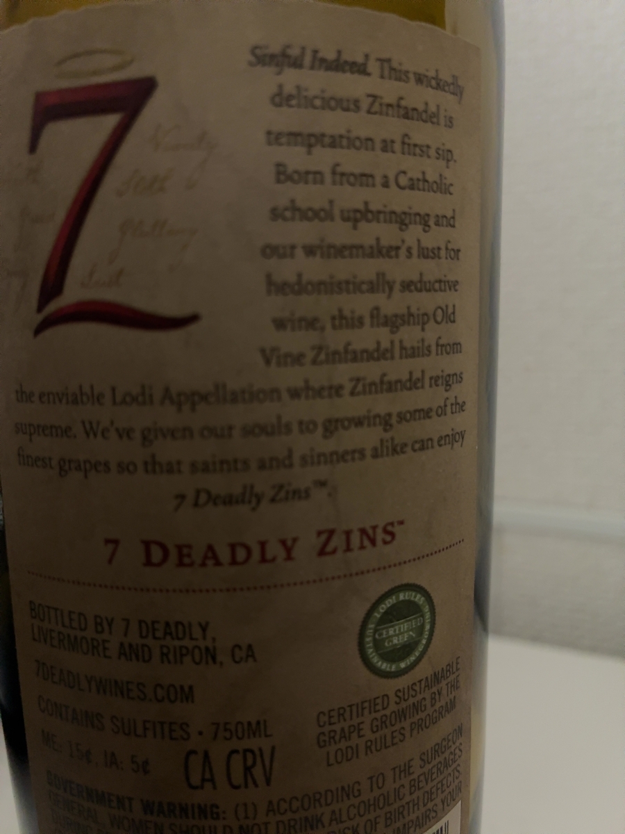 ラベルの大部分を占めるのは、ワインの紹介文です。上部には「Sinful Indeed. This wickedly delicious Zinfandel is temptation at first sip. Born from a Catholic school upbringing and our winemaker's lust for hedonistically seductive wine, this flagship Old Vine Zinfandel hails from the enviable Lodi Appellation where Zinfandel reigns supreme. We've given our souls to growing some of the finest grapes so that saints and sinners alike can enjoy 7 Deadly Zins™.」と書かれています。その下には、赤字で大きく「7 DEADLY ZINS」と記されており、その下には「BOTTLED BY 7 DEADLY, LIVERMORE AND RIPON, CA」と製造元と所在地が記載されています。さらにその下には「7DEADLYWINES.COM」というウェブサイトのアドレスがあります。 ラベルの下部には、ワインに関する情報がいくつか書かれています。「CONTAINS SULFITES - 750ML」とあり、亜硫酸塩が含まれていることと内容量が示されています。その右隣には「CA CRV」と「ME: 15¢, IA: 5¢」という文字が見えます。 ラベルの右下には、円形のロゴがあり、「LODI RULES CERTIFIED GREEN SUSTAINABLE WINEGROWING」と書かれています。その下には、「CERTIFIED SUSTAINABLE GRAPE GROWING BY THE LODI RULES PROGRAM」と、ロディ・ルールズ・プログラムによる持続可能なブドウ栽培の認証を受けていることが明記されています。