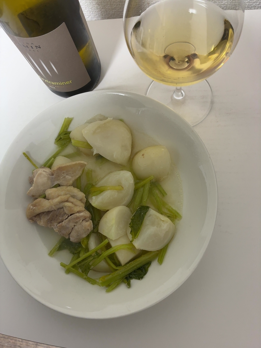 白いテーブルの上に、白い器に入った料理と白ワインが置かれています。 画像の中央には、白い器に入った料理が写っています。料理は、鶏肉とカブ、カブの葉を煮込んだもののようです。鶏肉は左側に寄せて盛り付けられており、カブは皮をむいて一口大に切られたものが複数個、カブの葉は緑色の茎の部分が目立つように添えられています。全体的に薄い色の出汁で煮込まれているように見えます。 料理の右奥には、脚付きのワイングラスに注がれた白ワインがあります。ワインは透明感のある淡い黄色をしており、グラスの表面には水滴のようなものが付着しています。 ワイングラスの左奥には、緑色のガラス瓶に入ったワインボトルが置かれています。ボトルのラベルは茶色で、上部に「INA」と読める文字があり、その下に「AMIN」と続く文字が見えます。さらにその下には「urztraminer」と書かれており、その下には「ATO ADIGE DOC」と「2024」という文字が確認できます。