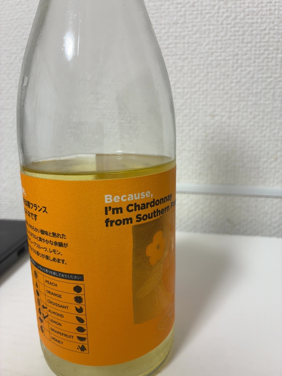 この画像には、オレンジ色のラベルが貼られたワインボトルが写っています。 ラベルには、「Because, I'm Chardonnay from Southern Fr」と書かれています。また、日本語で「南フランス」という文字も見えます。その下には、ワインの風味を表すような様々な香りのリストがあり、「PEACH」「ORANGE」「CROISSANT」「ALMOND」「LEMON」「GRAPEFRUIT」「HONEY」といった言葉が、それぞれの香りを象徴するイラストと共に並んでいます。ワインの色は淡い黄色で、ガラスボトル越しに見えます。背景は白い壁で、少しぼやけています。
