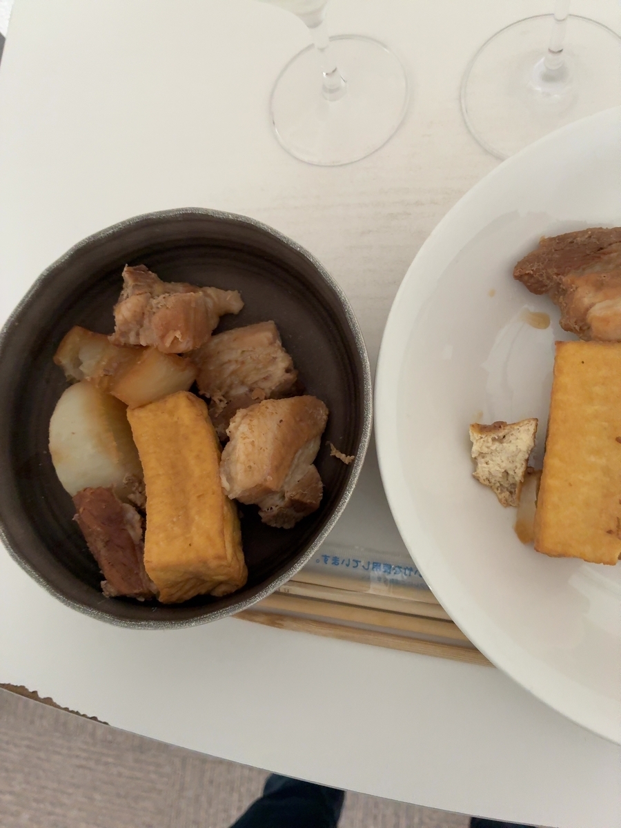 お皿の左側には、黒い器に入った料理が見えます。器の中には、角切りにされた豚肉や大根、そして厚揚げのようなものが盛り付けられています。豚肉は、脂身と赤身のコントラストが食欲をそそるような色合いをしています。大根は、白い肌が上品な印象を与えています。厚揚げは、黄金色に揚がっており、香ばしさが感じられそうです。 お皿の右側には、白い大きなお皿が置かれています。こちらにも、豚肉と厚揚げのようなものが盛り付けられています。豚肉は、先ほどのものと同じように、脂身と赤身が見て取れます。厚揚げは、先ほどのものよりも少し小さめのサイズで、こちらも美味しそうです。 お皿の下には、割り箸が2膳置かれています。箸袋には、青い文字で何らかの文字が印刷されていますが、残念ながらはっきりと読み取ることはできません。 背景には、2つのワイングラスが置かれています。グラスは透明で、光を反射してキラキラと輝いています。まるで、これから始まる食事への期待感を高めているかのようです。全体的に、落ち着いた雰囲気の中で、美味しい料理とお酒を楽しむひとときが想像されます。