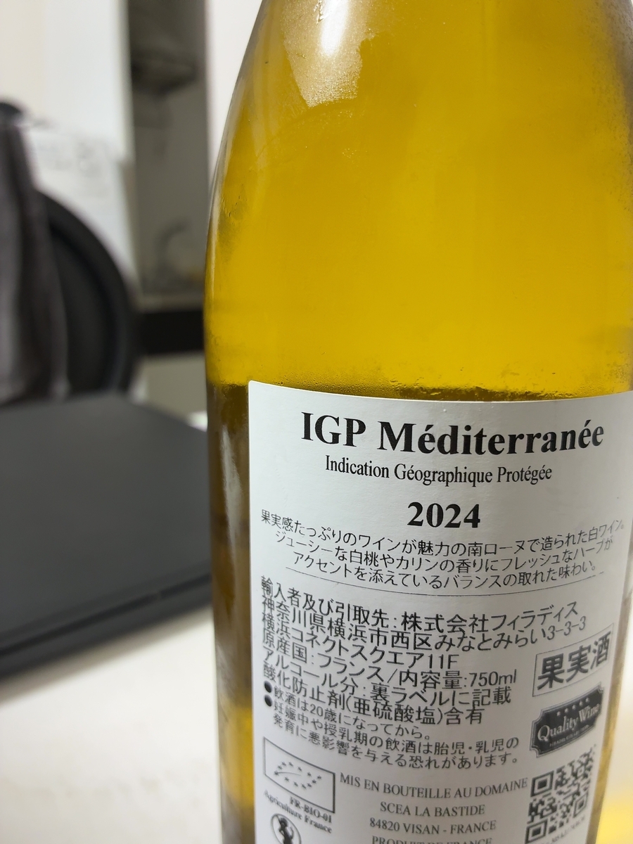 この画像は、ワインボトルのラベル部分を捉えたものです。 ラベルには「IGP Méditerranée」と書かれており、その下に「Indication Géographique Protégée」とフランス語で地理的表示保護の記載があります。さらに「2024」という数字が大きく表示されています。その下には、ワインの味わいに関する説明が日本語で書かれており、「果実感たっぷりのワインが魅力の南ローヌで造られた白ワイン。ジューシーな白桃やカリンの香りにフレッシュなハーブがアクセントを添えているバランスの取れた味わい。」と記されています。 次に、輸入者と引取先に関する情報があり、「輸入者及び引取先:株式会社フィラディス 神奈川県横浜市西区みなとみらい3-3-3 横浜コネクトスクエア11F」と記載されています。その下には「原産国:フランス/内容量:750ml」とあり、右側には「果実酒」という文字が見えます。さらに「アルコール分:裏ラベルに記載 酸化防止剤(亜硫酸塩)含有」と続き、その下には「飲酒は20歳になってから。」と「妊娠中や授乳期の飲酒は胎児・乳児の発育に悪影響を与える恐れがあります。」という注意書きが書かれています。 ラベルの左下には、有機認証マークと思われる「FR-BIO-01 Agriculture France」の文字とロゴが確認できます。右下には「MIS EN BOUTEILLE AU DOMAINE SCEA LA BASTIDE 84820 VISAN - FRANCE」というフランス語の記載があり、その隣にはQRコードが複数配置されています。ボトルの色は鮮やかな黄色で、中身のワインの色を反映しているようです。ボトルの表面には結露のような水滴がついており、冷えている様子がうかがえます。