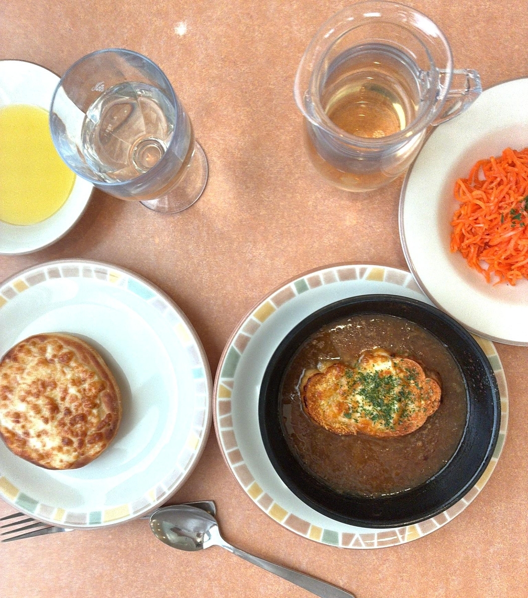 テーブルに置かれた食事の様子が、上から捉えられています。 画像の中央には、茶色いスープが入った黒い器があり、その中にはチーズが溶けて焦げ目がついたパンが浮かんでいます。パンの上には緑色のパセリが散らされています。この器は、白地に淡い色合いの四角い模様が縁に描かれたお皿の上に置かれています。その左隣には、同じ模様のお皿に、丸くて平たいパンが置かれています。このパンも表面に焦げ目がついていて、チーズが溶けているように見えます。 右上の白いお皿には、細かく刻まれたオレンジ色の人参が山盛りに盛られており、その上にも緑色のパセリが散らされています。その隣には、透明なガラスのピッチャーに琥珀色の液体が入っています。左上には、透明なワイングラスに水が注がれており、その隣の白い小皿には、黄色い液体が入っています。 テーブルは茶色で、少しざらざらとした質感に見えます。全体的に、温かみのある食卓の雰囲気が伝わってきます。
