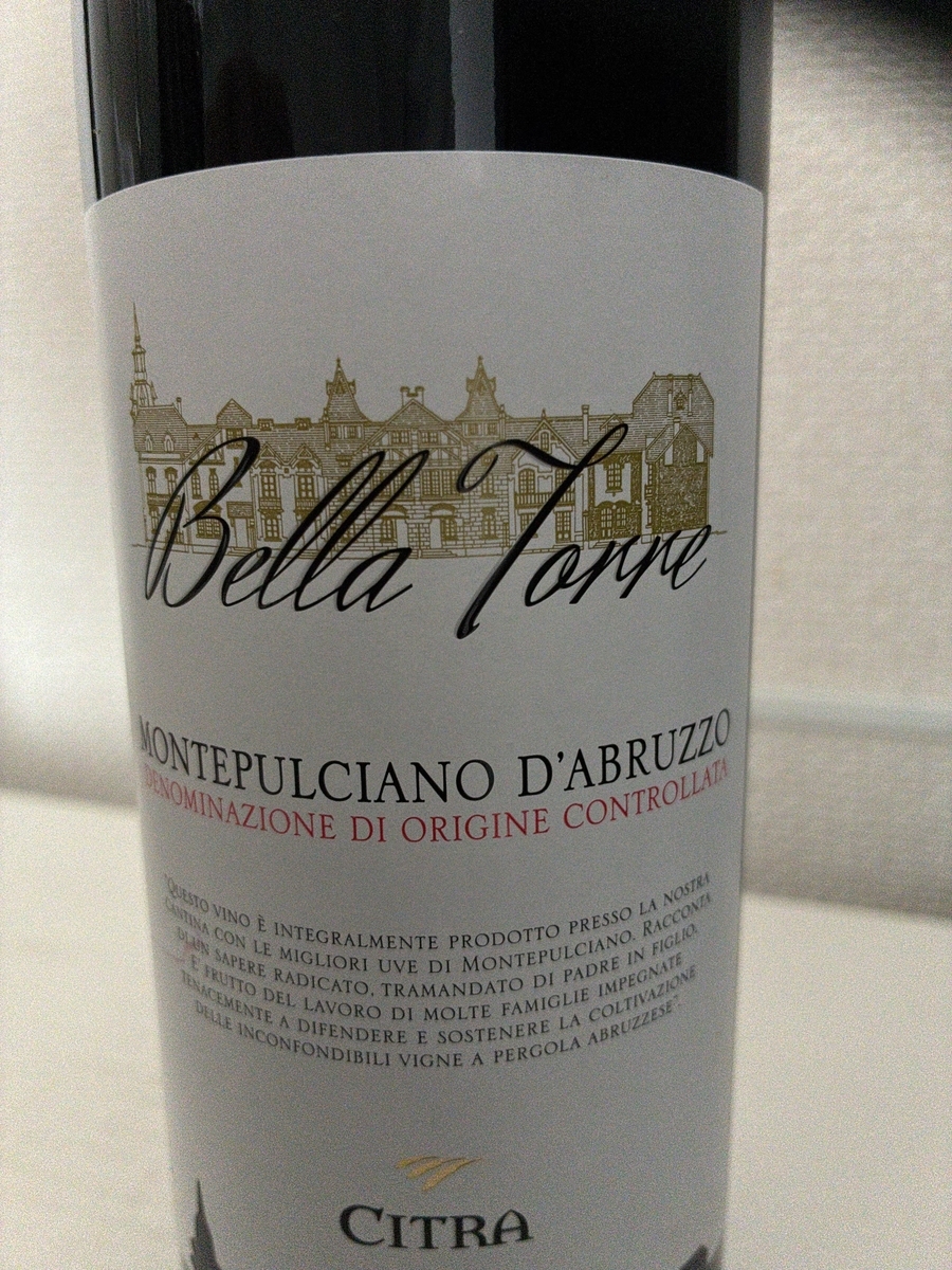 ラベルの中央には「Bella Torre」と書かれた筆記体のロゴがあり、その背景には金色の線で描かれたヨーロッパ風の建物が広がっています。ロゴの下には「MONTEPULCIANO D'ABRUZZO」と黒い大文字で書かれており、さらにその下には赤字で「DENOMINAZIONE DI ORIGINE CONTROLLATA」と記されています。 その下には、このワインに関する説明文がイタリア語で書かれています。読み取れる範囲では、「QUESTO VINO È INTEGRALMENTE PRODOTTO PRESSO LA NOSTRA CANTINA CON LE MIGLIORI UVE DI MONTEPULCIANO. RACCONTA DI UN SAPERE RADICATO, TRAMANDATO DI PADRE IN FIGLIO E FRUTTO DEL LAVORO DI MOLTE FAMIGLIE IMPEGNATE TENACEMENTE A DIFENDERE E SOSTENERE LA COLTIVAZIONE DELLE INCONFONDIBILI VIGNE A PERGOLA ABRUZZESE」と書かれています。 ラベルの一番下には「CITRA」という文字が黒い大文字で書かれており、その上には金色の小さなロゴマークが見えます。ラベル全体はクリーム色で、わずかにざらつきのある紙のような質感が感じられます。ボトルは黒いガラス製で、ラベルの上下でその色が確認できます。