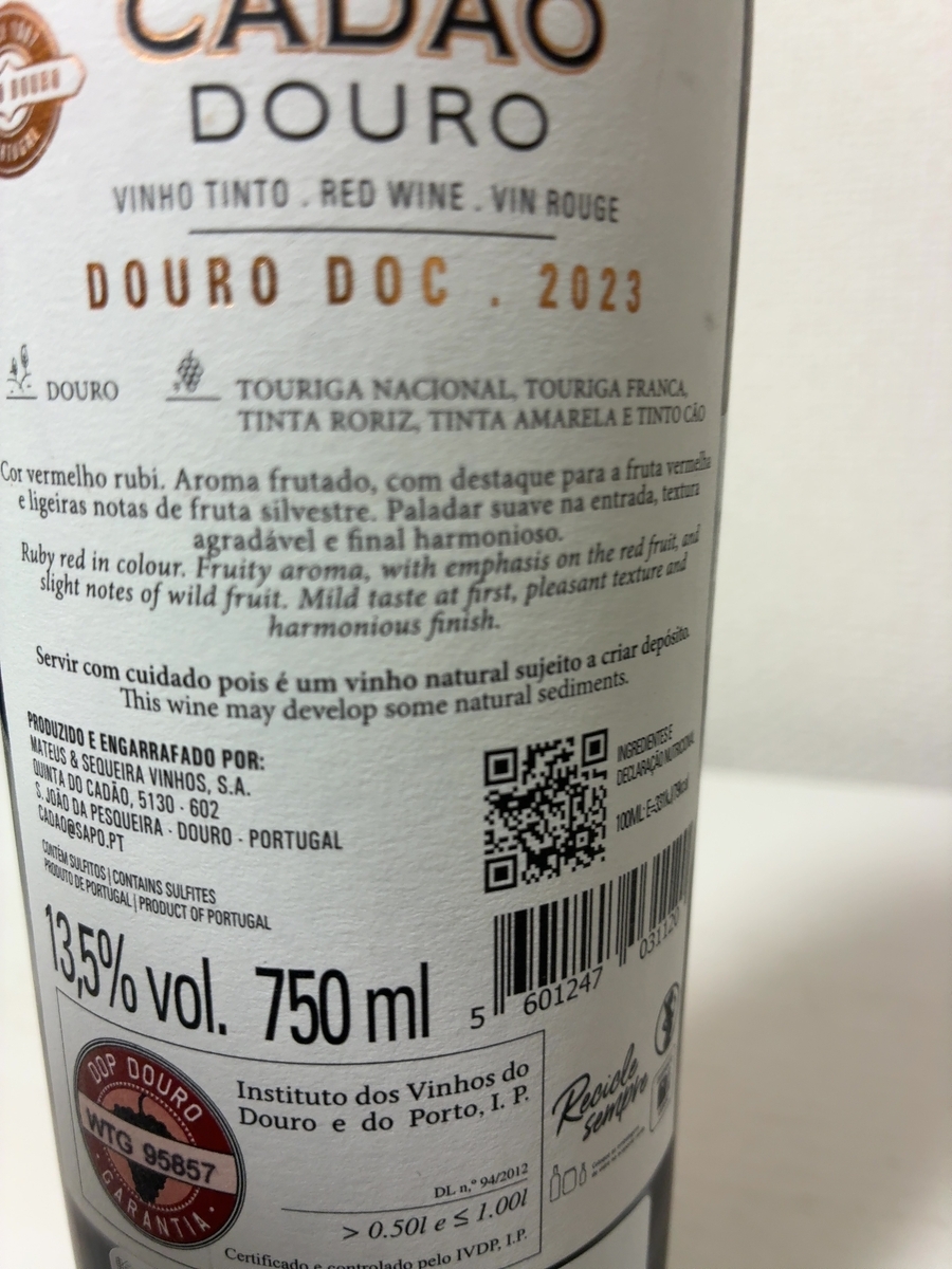 この画像は、ワインボトルのラベルを写したものです。 ラベルの上部には「CADAO DOURO」と大きく書かれており、その下には「VINHO TINTO・RED WINE・VIN ROUGE」と、ポルトガル語、英語、フランス語で赤ワインであることが示されています。さらにその下には「DOURO DOC. 2023」と、ドウロDOC(原産地呼称)とヴィンテージが記載されています。 ラベルの中央部分には、ブドウの品種として「TOURIGA NACIONAL, TOURIGA FRANCA, TINTA RORIZ, TINTA AMARELA E TINTO CÃO」が挙げられています。その下には、ワインのテイスティングノートがポルトガル語と英語で書かれています。ポルトガル語では「Cor vermelho rubi. Aroma frutado, com destaque para a fruta vermelha e ligeiras notas de fruta silvestre. Paladar suave na entrada, textura agradável e final harmonioso.」とあり、英語では「Ruby red in colour. Fruity aroma, with emphasis on the red fruit, and slight notes of wild fruit. Mild taste at first, pleasant texture and harmonious finish.」と書かれています。これは、ルビーレッドの色合いで、赤果実と野生の果実の香りが特徴的で、口当たりは滑らかで心地よいテクスチャーと調和の取れた余韻があることを示しています。 さらに下には、「Servir com cuidado pois é um vinho natural sujeito a criar depósito. This wine may develop some natural sediments.」と、自然な沈殿物が生じる可能性があるため、注意して提供するよう促す注意書きがポルトガル語と英語で書かれています。