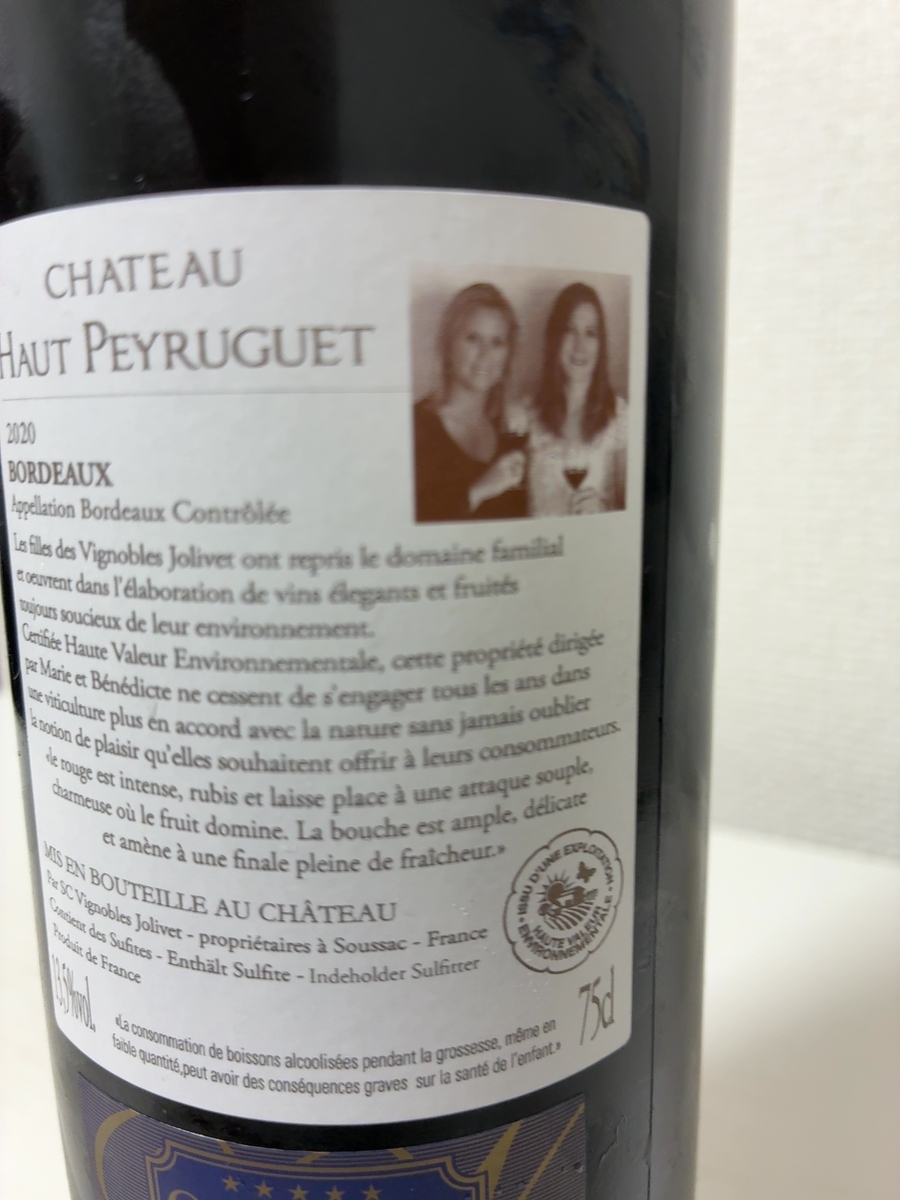 ラベルの上部には「CHATEAU HAUT PEYRUGUET」と書かれており、その下には「2020 BORDEAUX Appellation Bordeaux Contrôlée」と続いています。ラベルの右側には、2人の女性の顔写真が配置されています。 写真の下には、ワインの生産者に関する説明文が書かれています。読み取れる範囲では、「Les filles des Vignobles Jolivet ont repris le domaine familial et oeuvrent dans l'élaboration de vins élégants et fruités toujours soucieux de leur environnement. Certifiée Haute Valeur Environnementale, cette propriété dirigée par Marie et Bénédicte ne cessent de s'engager tous les ans dans une viticulture plus en accord avec la nature sans jamais oublier la notion de plaisir qu'elles souhaitent offrir à leurs consommateurs. Ce rouge est intense, rubis et laisse place à une attaque souple, charmeuse où le fruit domine. La bouche est ample, délicate et amène à une finale pleine de fraîcheur.」と記されています。 さらにその下には、「MIS EN BOUTEILLE AU CHATEAU Par SC Vignobles Jolivet - propriétaires à Soussac - France Contient des Sulfites - Enthält Sulfite - Indeholder Sulfitter Produit de France」と書かれています。 ラベルの右下には、丸いロゴマークがあり、その中には蝶と畑のような絵柄が描かれ、「ISSU D'UNE EXPLOITATION HAUTE VALEUR ENVIRONNEMENTALE」という文字が確認できます。その右隣には「75cl」と容量が示されています。 ラベルの最下部には、妊娠中のアルコール摂取に関する注意書きがフランス語で書かれており、「«La consommation de boissons alcoolisées pendant la grossesse, même en faible quantité, peut avoir des conséquences graves sur la santé de l'enfant.»」と読み取れます。