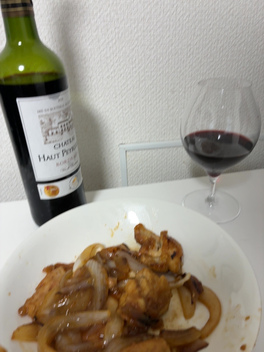 ワインボトルとグラス、そして料理が写し出された食卓の様子を捉えています。 画像の中央左には、緑色のガラス瓶に入ったワインボトルが置かれています。ボトルのラベルには「CHATEAU HAUT PEYRAUD」と書かれており、その上には城のような建物のイラストが描かれています。ボトルの右側には、ワイングラスが置かれており、グラスの中には赤ワインが注がれています。グラスの奥には、白い壁が見え、その壁には細かな凹凸のある模様が施されています。壁の手前には、白い細い棒状のものが横に伸びており、その下には白い四角い枠のようなものが見えます。画像の手前には、白い皿に盛られた料理が写っています。料理は、鶏肉と玉ねぎを甘辛いタレで炒めたようなもので、照りがあり、食欲をそそるような見た目をしています。