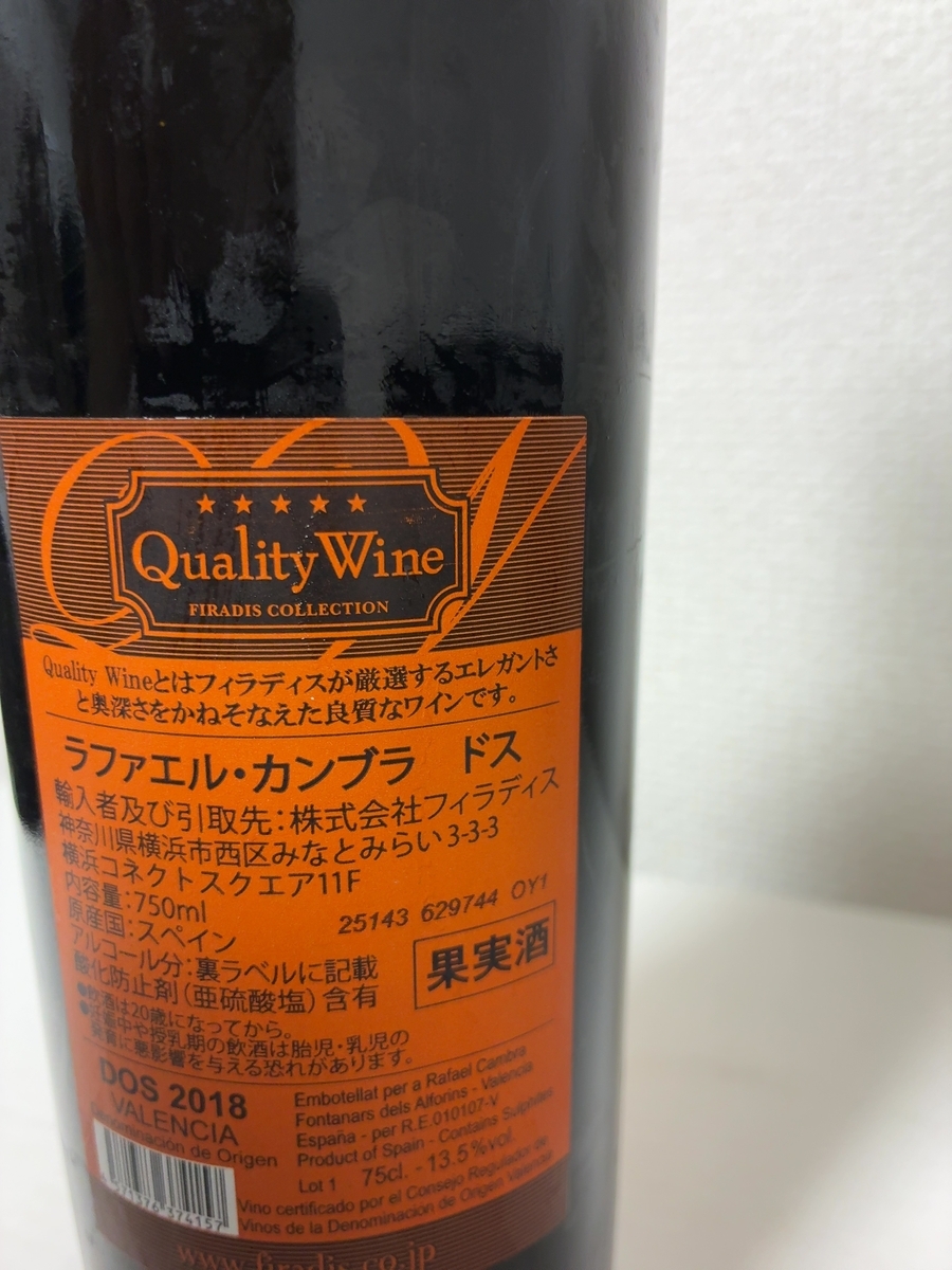 ラベルはオレンジ色を基調とし、上部には「Quality Wine」という文字と5つの星がデザインされています。その下には「FIRADIS COLLECTION」と書かれています。 ラベルの中央には、黒い文字で「Quality Wineとはフィラディスが厳選するエレガントさと奥深さをかねそなえた良質なワインです。」という説明文があります。その下には、大きな黒い文字で「ラファエル・カンブラ ドス」と商品名が書かれています。 さらにその下には、輸入者と引取先に関する情報が記載されています。「輸入者及び引取先:株式会社フィラディス 神奈川県横浜市西区みなとみらい3-3-3 横浜コネクトスクエア11F」とあります。 内容量は「750ml」、原産国は「スペイン」と明記されており、その右隣には「25143 629744 OY1」という数字と文字の組み合わせがあります。 アルコール分については「裏ラベルに記載」とあり、その横には「果実酒」と書かれた四角い枠があります。 酸化防止剤として「亜硫酸塩」が含まれていることが示されており、その下には「●飲酒は20歳になってから。」「●妊娠中や授乳期の飲酒は胎児・乳児の発育に悪影響を与える恐れがあります。」という注意書きが記載されています。 ラベルの下部には「DOS 2018 VALENCIA」とあり、その右側には英語でワインに関する詳細情報が書かれています。具体的には、「Embotellat per a Rafael Cambra Fontanars dels Alforins - Valencia España - per R.E.010107-V Product of Spain - Contains Sulphites Lot 1 75cl. - 13.5%Vol Vino certificado por el Consejo Regulador de Vinos de la Denominacion de Origen Valencia」と読み取れます。