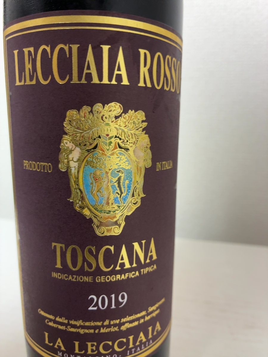 レッチャイア ロッソ 2019の表ラベルのクローズアップ。濃い赤紫色のラベルに金色の文字で「LECCIAIA ROSSO」と大きく書かれている。中央には金色の装飾枠に囲まれた紋章——青と金の盾形エンブレムを両脇から人物と植物のモチーフが支える彫刻的なデザイン。その下に「TOSCANA / INDICAZIONE GEOGRAFICA TIPICA」「2019」「LA LECCIAIA / MONTALCINO - ITALIA」と続く。白い背景に映えた、格調ある一本の佇まい。