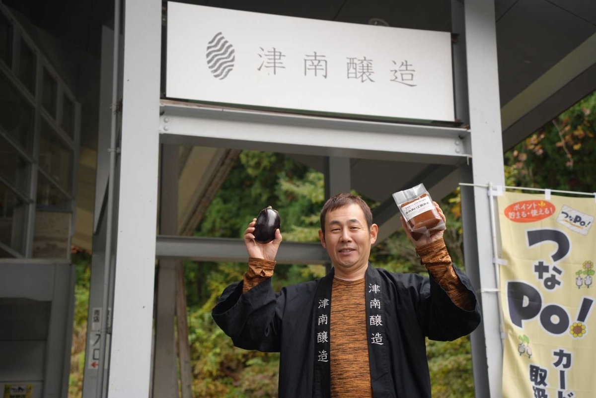 津南醸造の入口で担当者が丸なすと商品袋を掲げるスナップ—背後にロゴ看板と「つなPo!」の旗