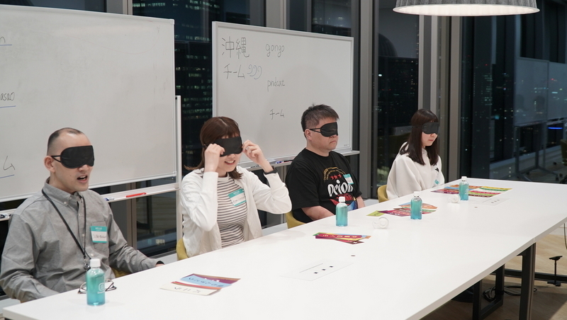長机に座る4人の社員。みんな黒いアイマスクをつけている。