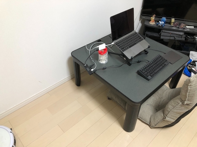 高さが低めの机と座椅子を使って仕事環境にしている。机は正方形でMacのノートPCとキーボードだけが置かれている。ノートPCをテレビに接続しており、変換器の位置がシェイカーのようなものでちょうどいい高さに調整されている。