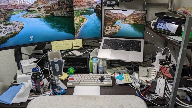 説明が難しいほどたくさんのものが置かれたデスク。大きなモニター2台とノートPC、キーボードがメイン。机の右側にラックがあり色々ものを収納している。大きなモニターには黄色い付箋が3枚貼られている。そのなかの1枚に「ゆっくりはなす リアクションは大きく」と書かれていてとてもよい。