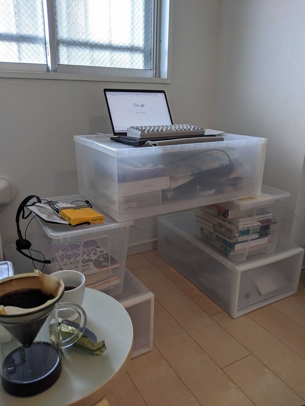 透明の衣装ケースを左右に2個、そのうえに1個置き、机として使用している。衣装ケースの上にノートPCが置かれ、衣装ケースの中にはWEB+DB PRESSをはじめたくさんの書籍が入っている。手前に小さな白い丸テーブルがありコーヒーが置かれている。