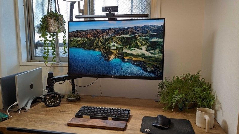 木目調の机に大きなモニターとキーボード、木材を使用したアームレストが置かれている。机の右奥には緑が置かれていて目に優しい。