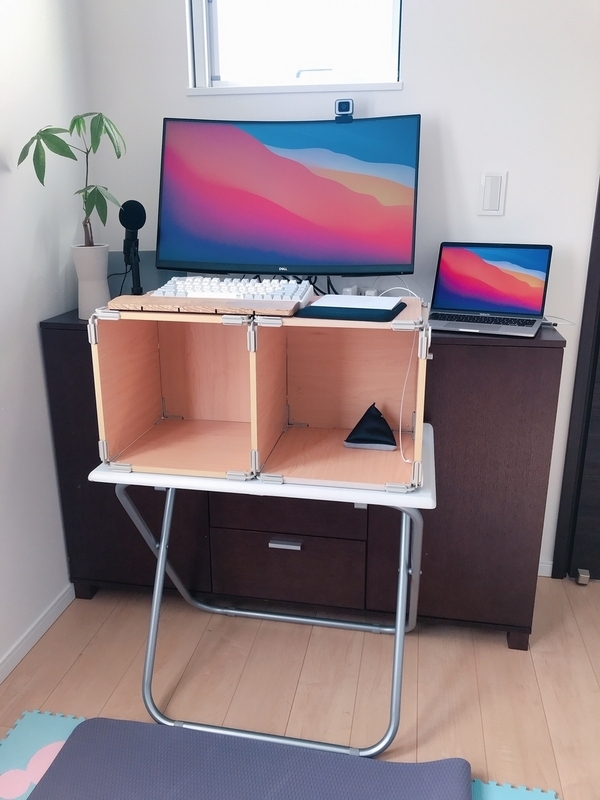 小さな台の上にカラーボックスが横向きに置かれていて、それを机として代用している。横向きカラーボックスの上に、モニターとキーボードとアームレストが置かれている。MacのノートPCはカラーボックスより奥の棚に置かれている。