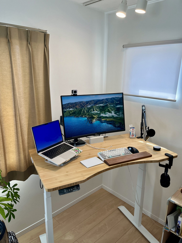 昇降式デスクにモニターとキーボードとアームレストが置かれている。右側にはアーム付きのマイクが置かれ、左側にMacのノートPCが置かれている。机は明るい色の木材を使用しておりクリーンな印象。